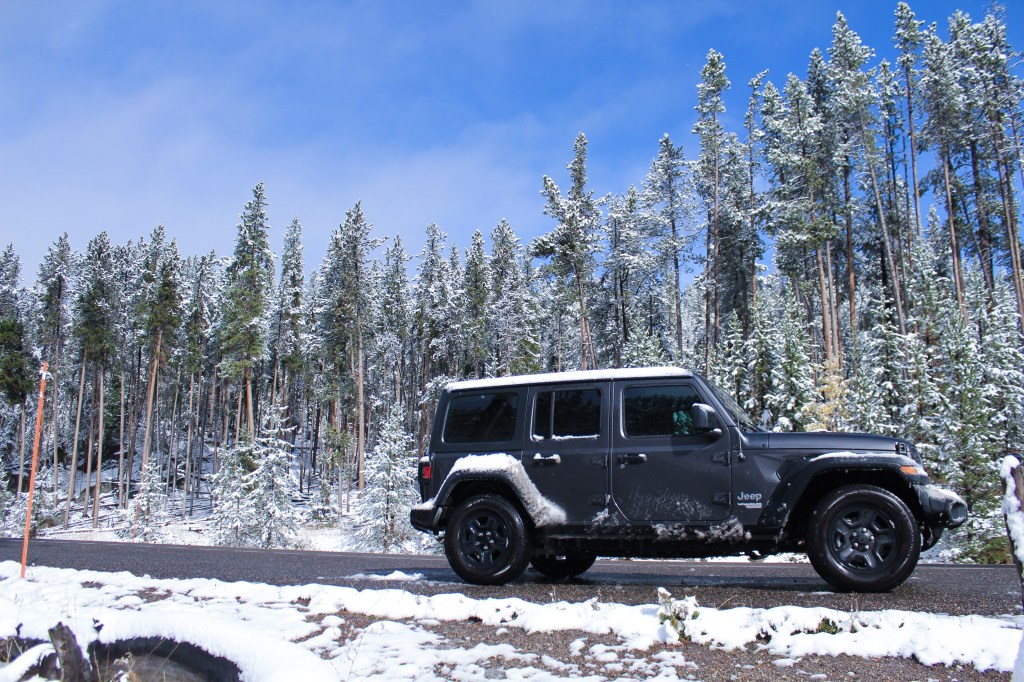 2018 Jeep Wrangler Unlimited Sport JL dark gray on a wet road with snow. There is a dusting of snow on the side of the roads and covering the tall evergreen trees in the background. Blue sky in the far background.