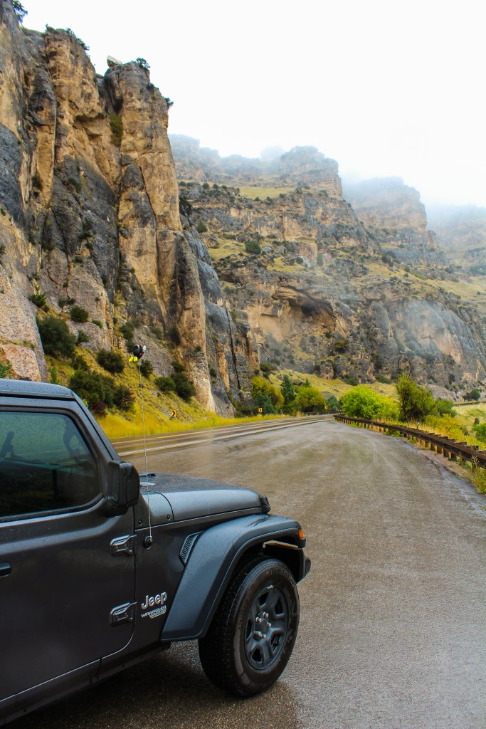The front half of a 2018 Jeep Wrangler Unlimited Sport on the left side of the image. In the background, the upward view of mountain cliffs of which the tops are covered in fog. 
